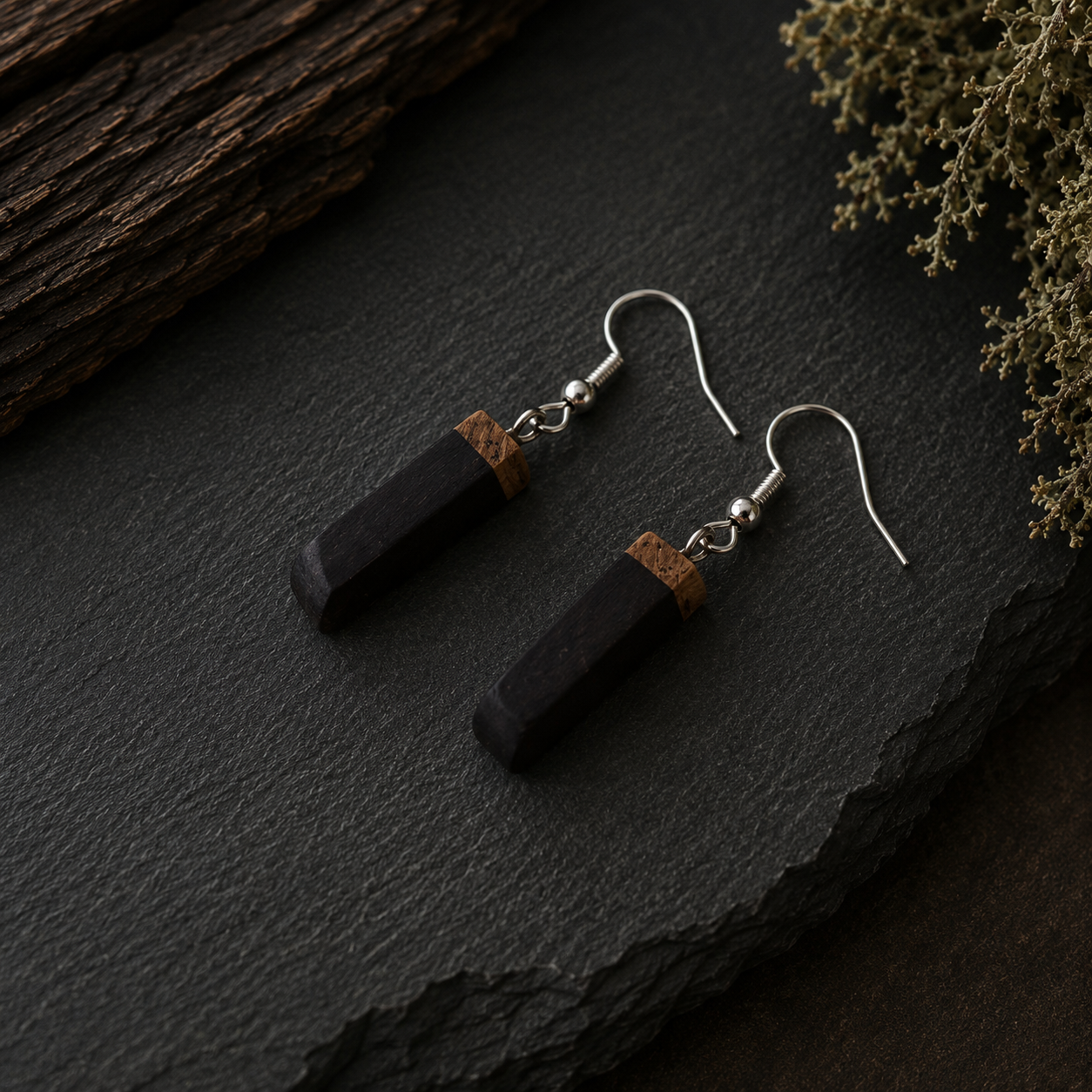 A pair of drop earrings with rectangular black pendants and small brown textured bands, hanging from silver fishhook wires on a dark slate surface with dried botanicals and wood in the background.