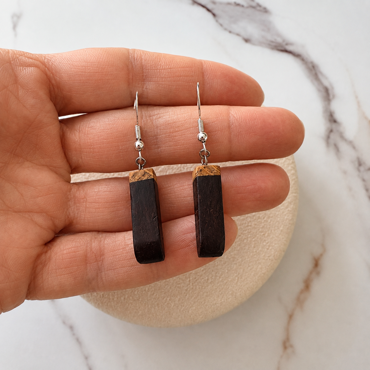 A pair of dark brown rectangular drop earrings with silver fishhook wires and small wooden caps, held between a person's fingers over a light marble surface.