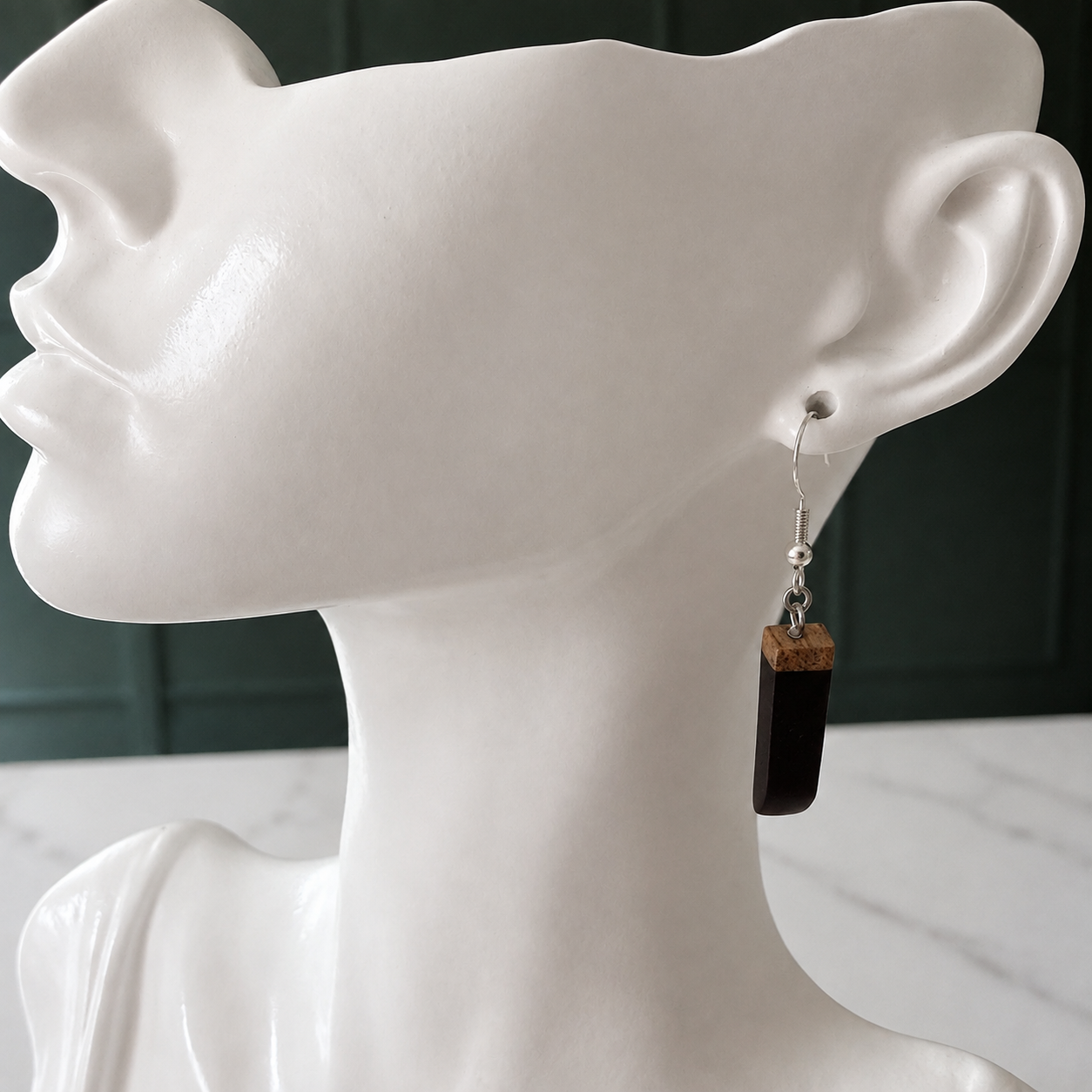 A single drop earring displayed on a white mannequin head. The earring features a silver fishhook wire, a small silver bead, and a dark brown wooden block with a glossy black stone or glass insert.