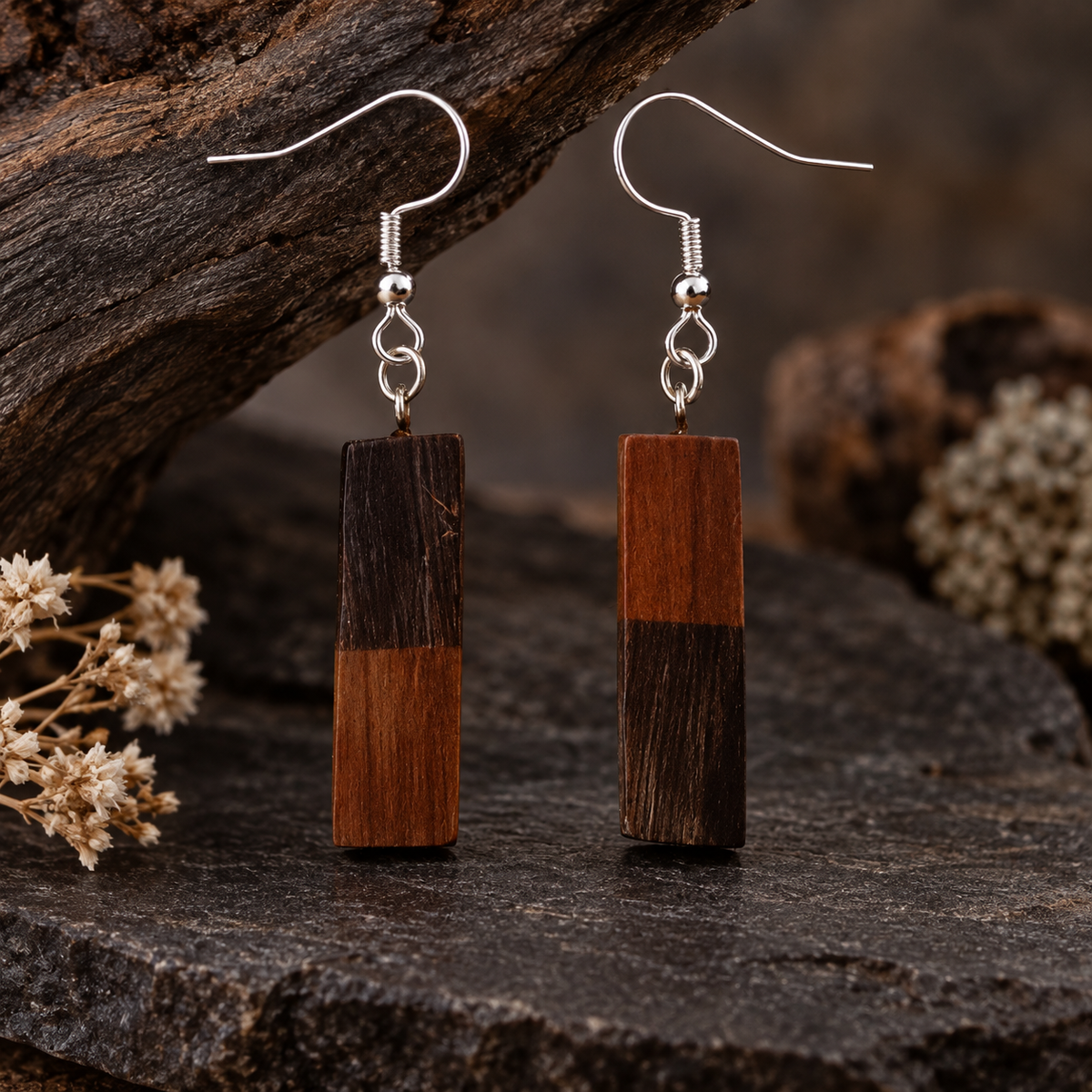 A pair of rectangular drop earrings made from two-tone wood blocks with silver fishhook wires, displayed on a dark textured stone surface with rustic wood and dried flowers in the background.