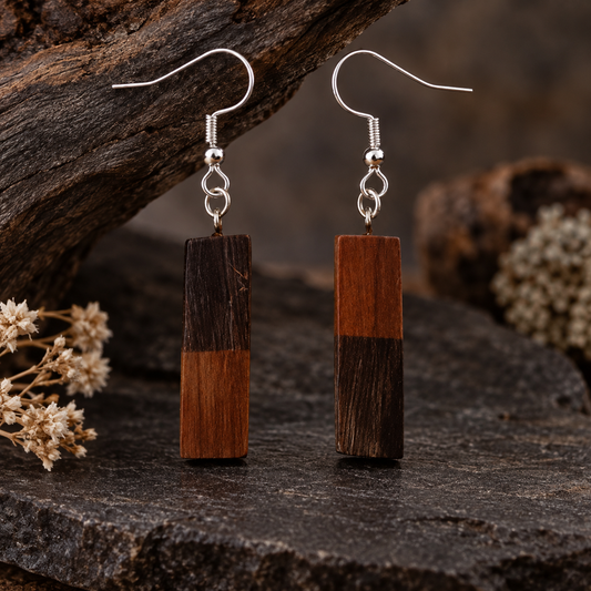 A pair of rectangular drop earrings made from two-tone wood blocks with silver fishhook wires, displayed on a dark textured stone surface with rustic wood and dried flowers in the background.