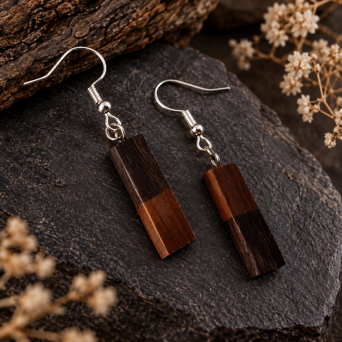 A pair of rectangular drop earrings with dark wood blocks in two brown tones and silver fishhook wires, displayed on a dark stone surface with dried flowers and a piece of wood in the background.