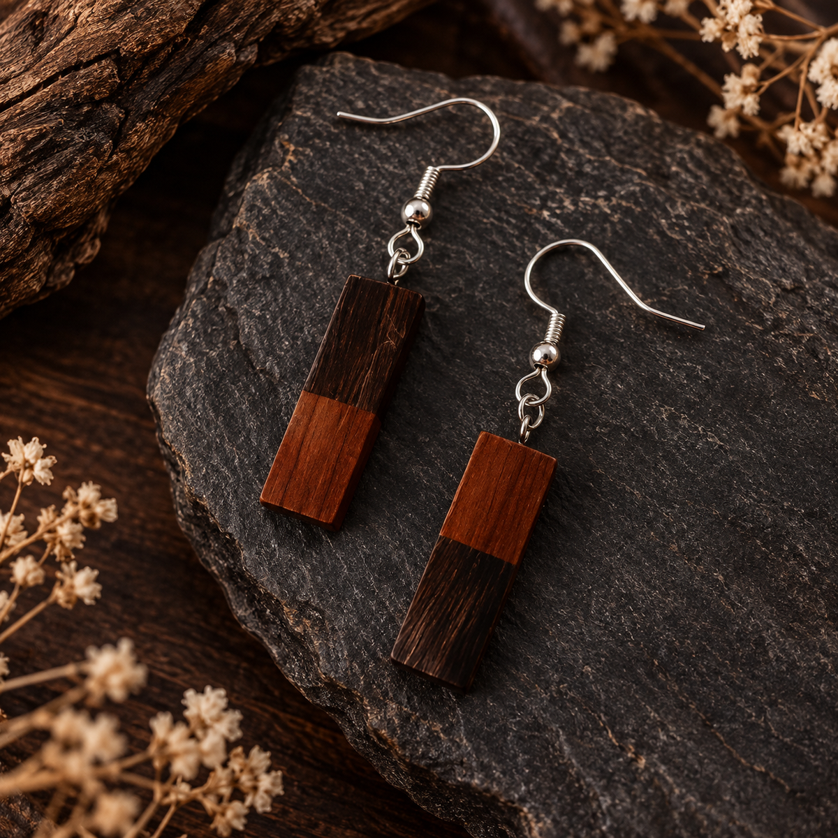 A pair of rectangular drop earrings with a two-tone wood design in dark brown and reddish-brown, suspended from silver fishhook wires, displayed on a dark textured rock with dried flowers.
