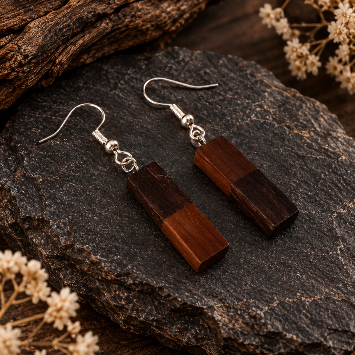 A pair of rectangular drop earrings made from two-tone dark brown wood blocks, suspended from silver fishhook wires, displayed on a dark textured rock with dried flowers.