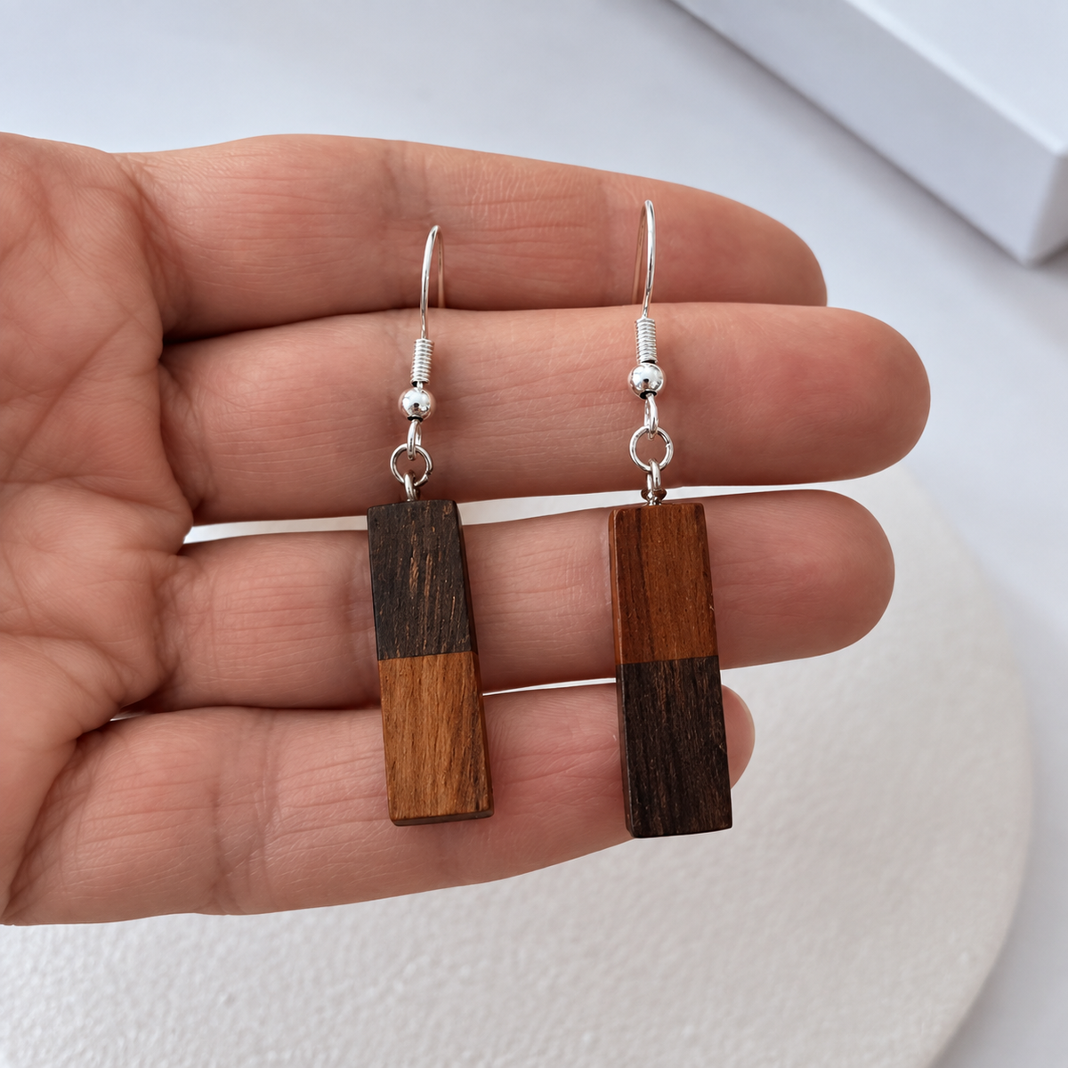 A pair of rectangular drop earrings with a two-tone wood design in dark and medium brown, hanging from silver fishhook wires, held in a person's hand.
