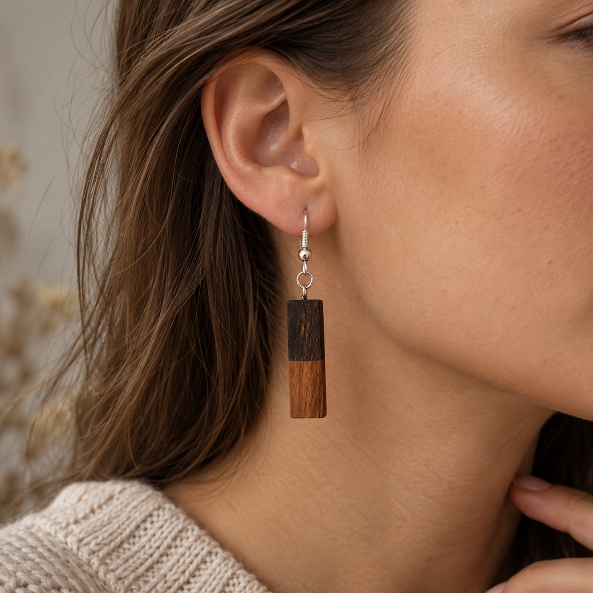 A close-up of a woman wearing a rectangular wooden dangle earring with a dark and light wood split design and a silver fishhook wire.