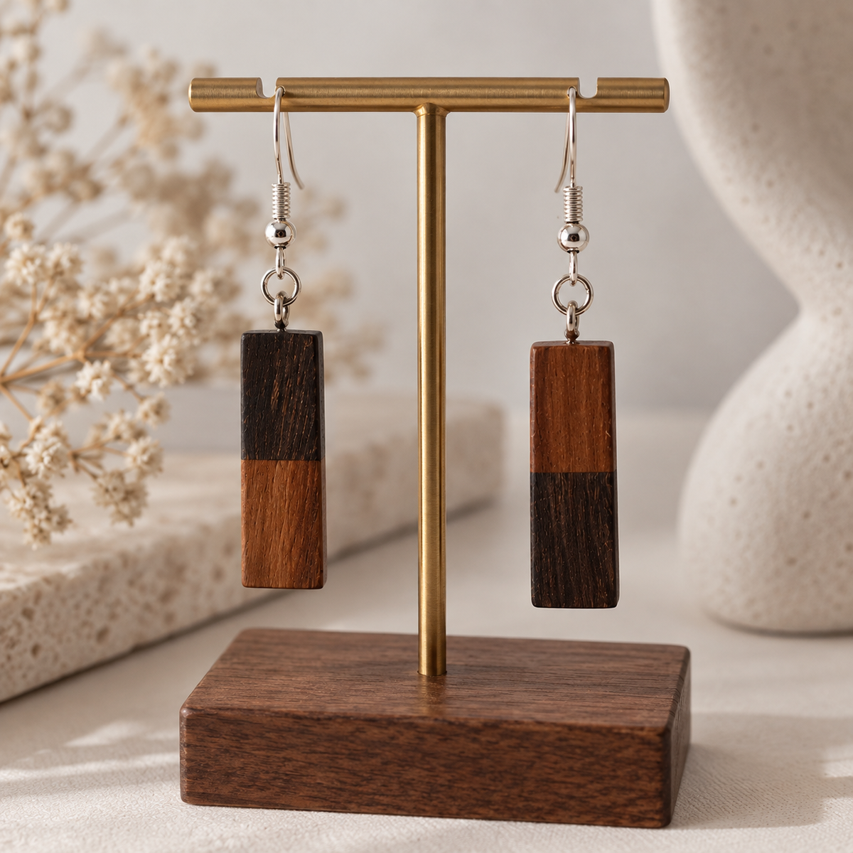 A pair of rectangular drop earrings with a two-tone wood finish hanging from a brass T-stand on a wooden base. The earrings feature a light brown wood top half and a dark brown wood bottom half, displayed against a softly blurred neutral background with dried flowers.