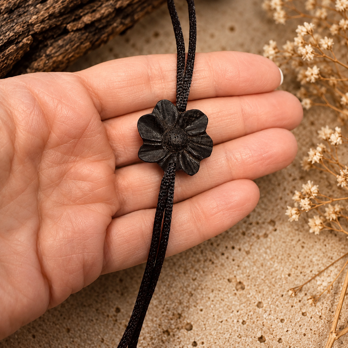 A black woven cord bracelet with a textured black flower charm, held in a person's hand against a sandy background with dried flowers and a piece of wood.