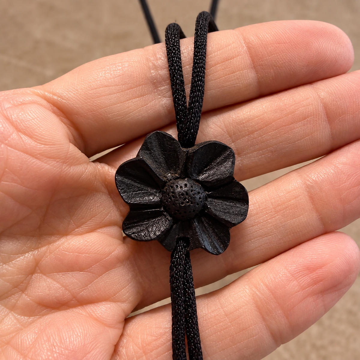A close-up of a hand holding a black flower-shaped pendant necklace on a black cord, with the textured flower center and woven cord clearly visible against the skin.
