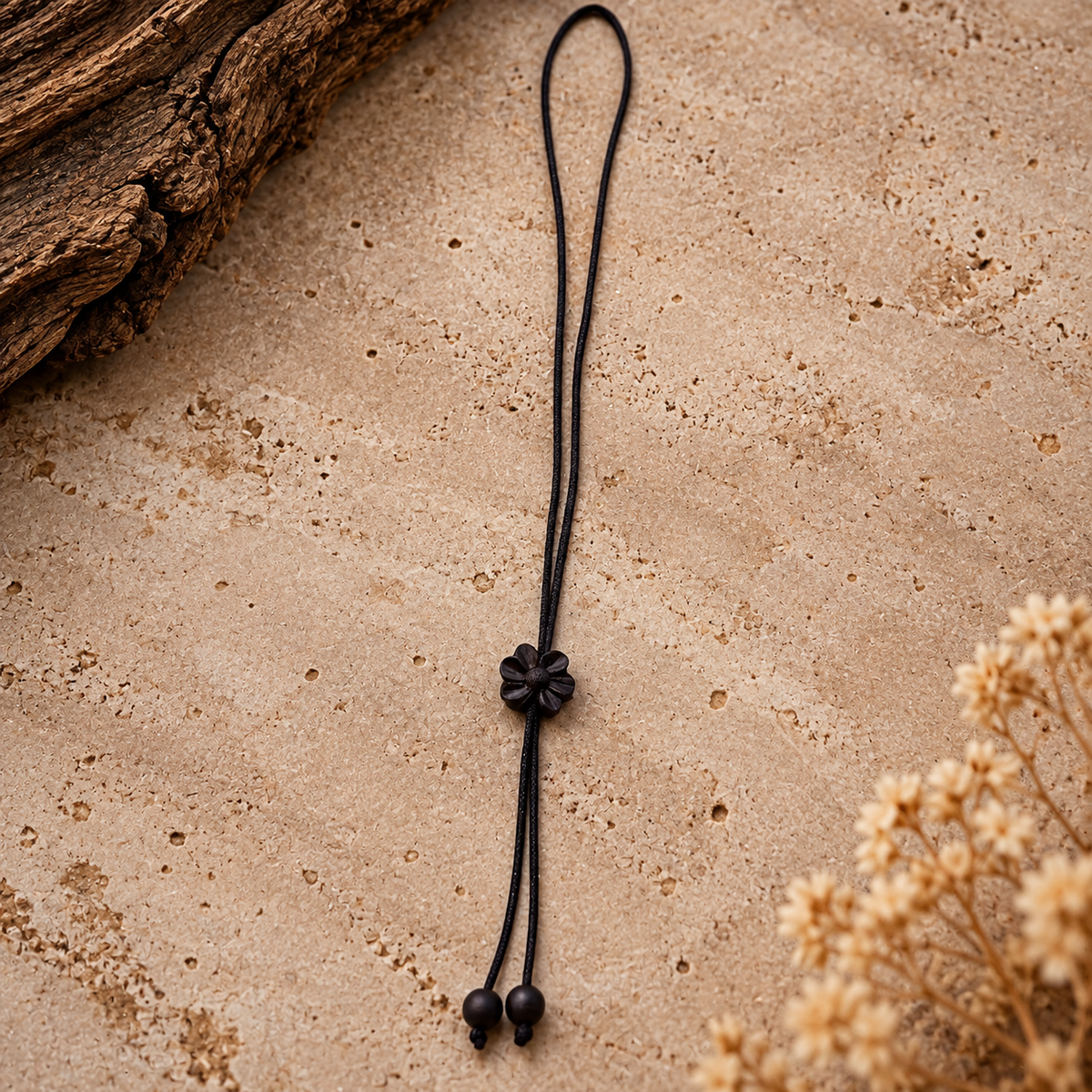 A black cord necklace with a flower-shaped centerpiece and two small beads at the ends, laid on a textured beige surface with dried flowers and a piece of wood in the corners.