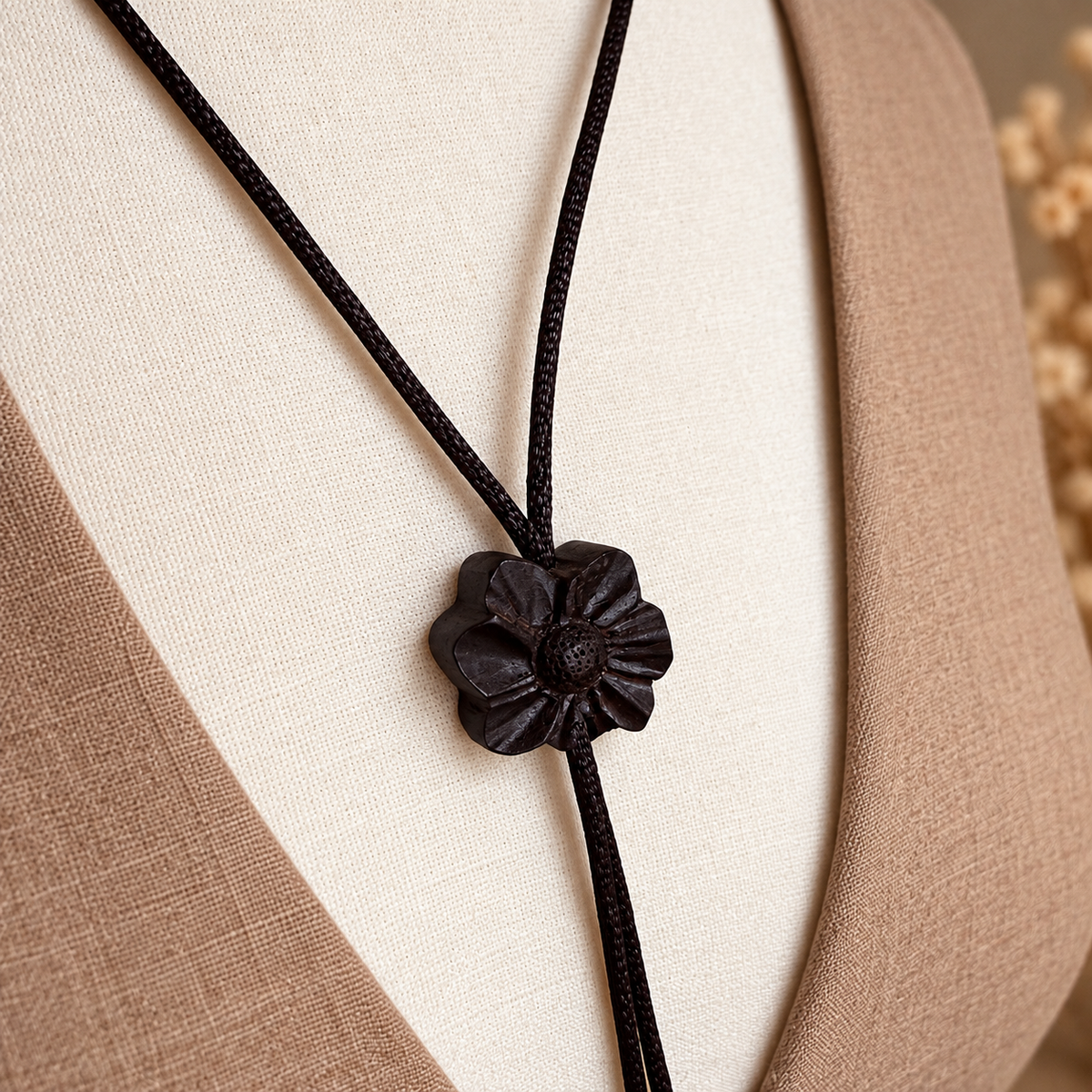 A close-up of a black cord necklace with a dark brown carved wooden flower pendant, displayed on a beige fabric mannequin bust.