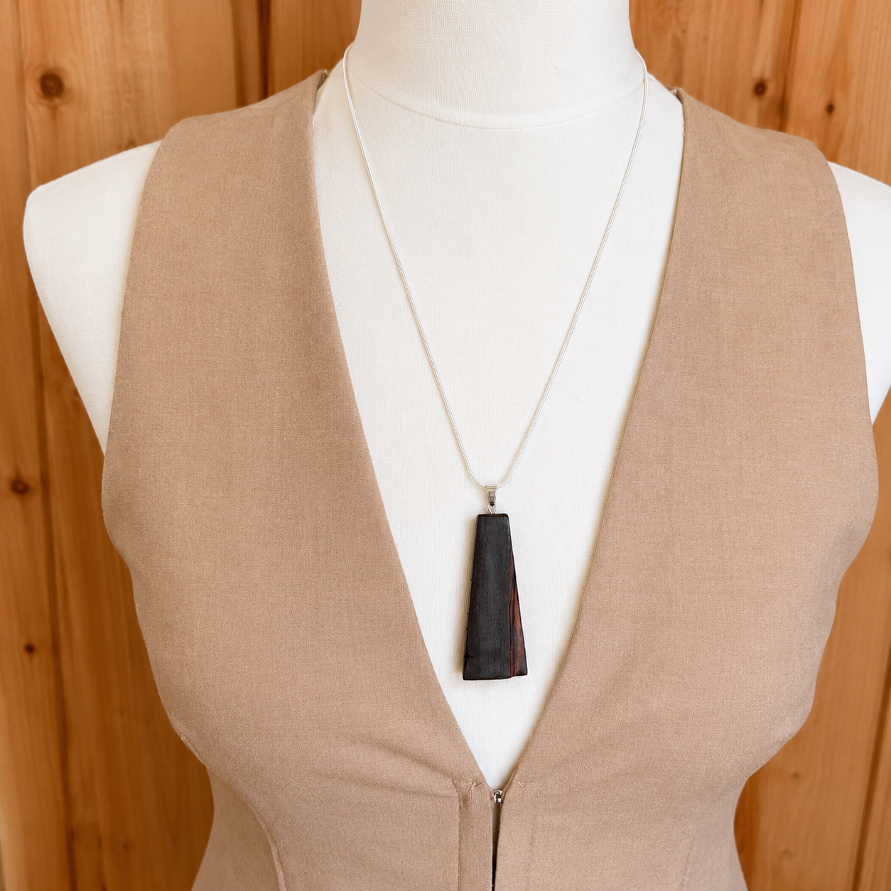 Mannequin wearing a beige vest with a necklace featuring a black pendant against a wooden background