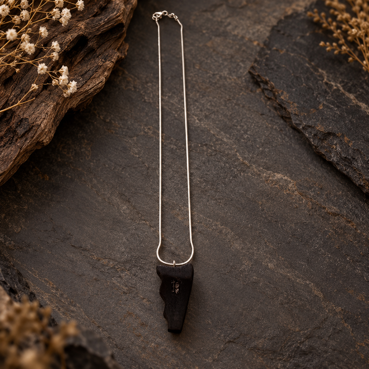 A silver chain necklace with a black stone pendant, displayed on a dark textured slate surface with dried flowers and wood accents.