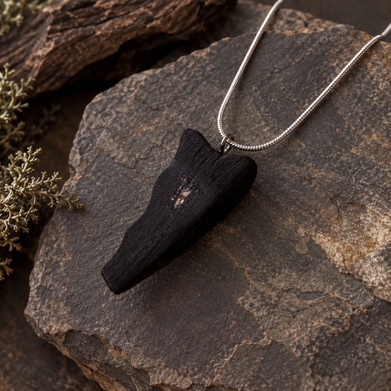 A black textured wood pendant necklace on a silver snake chain, displayed on a dark textured rock with dried botanicals.
