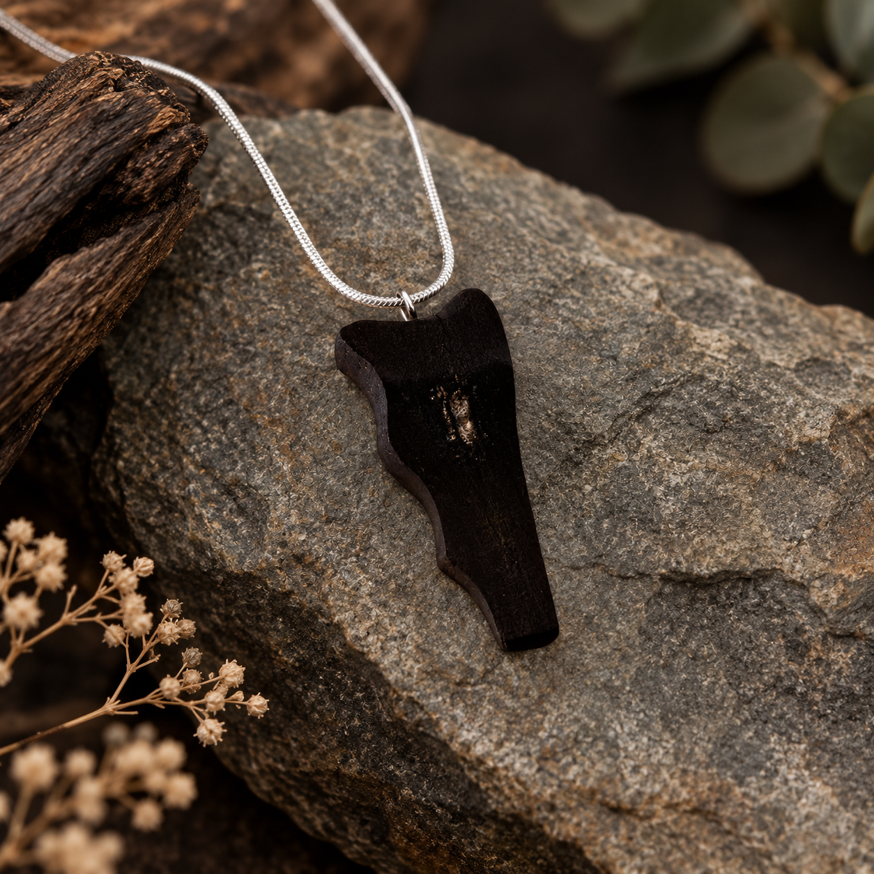 A dark brown wooden pendant necklace with a silver chain, displayed on a textured gray rock with dried flowers and rustic wood in the background.