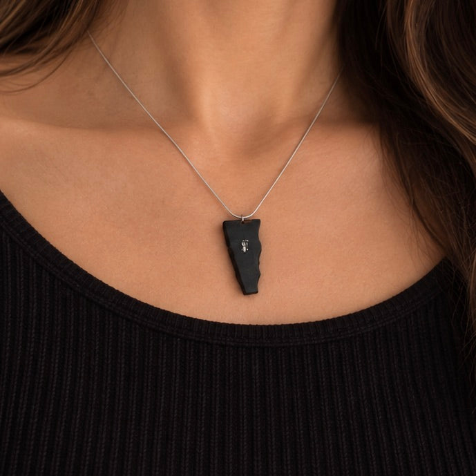 A close-up of a person's neck and chest wearing a silver chain necklace with a black irregularly shaped pendant featuring a small white monogram.