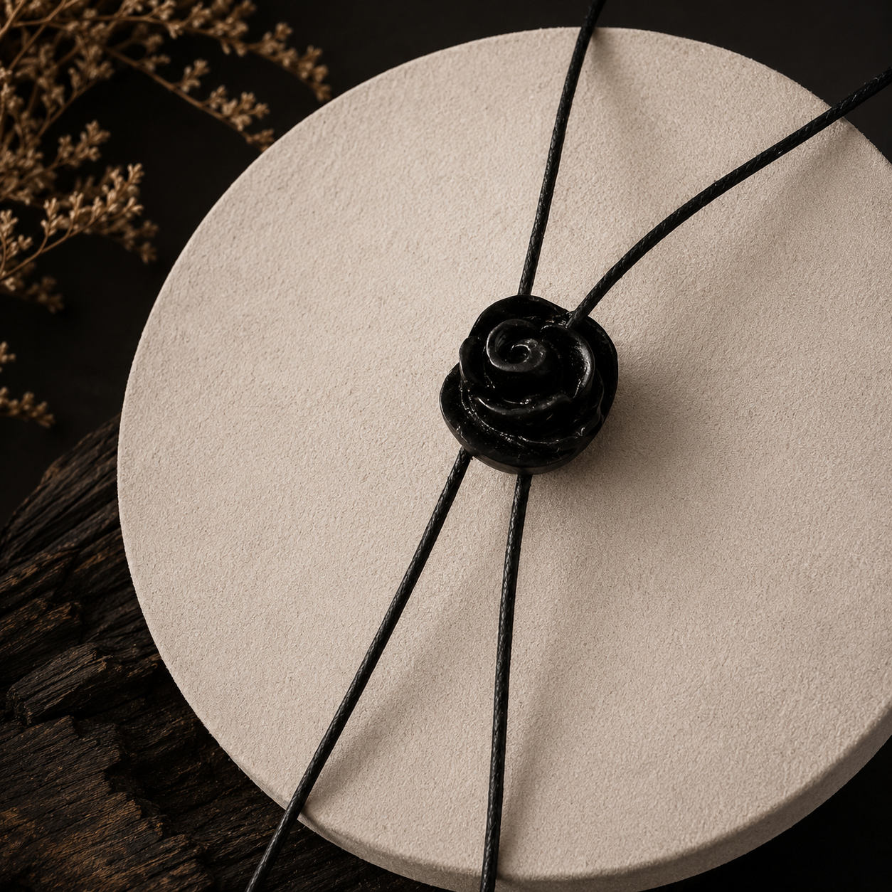 A black cord necklace with a glossy black rose-shaped pendant, displayed on a round beige textured stand with dark wood and dried flowers in the background.
