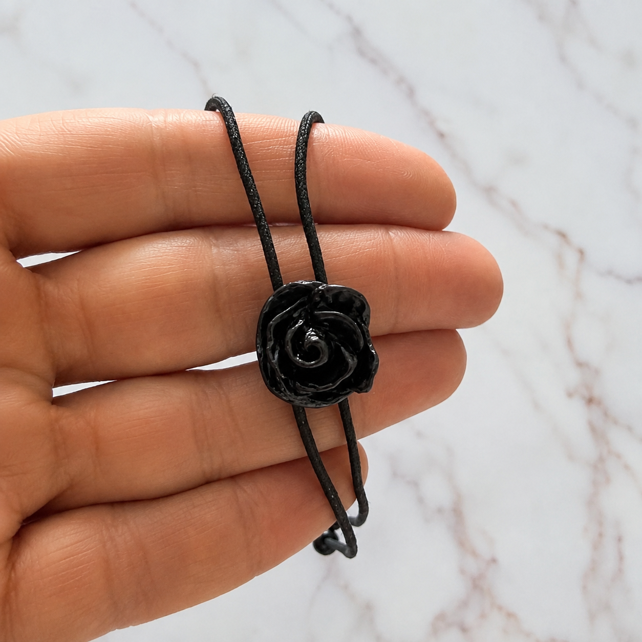 A hand holding a black adjustable cord bracelet with a glossy black rose-shaped charm, set against a white marble background.