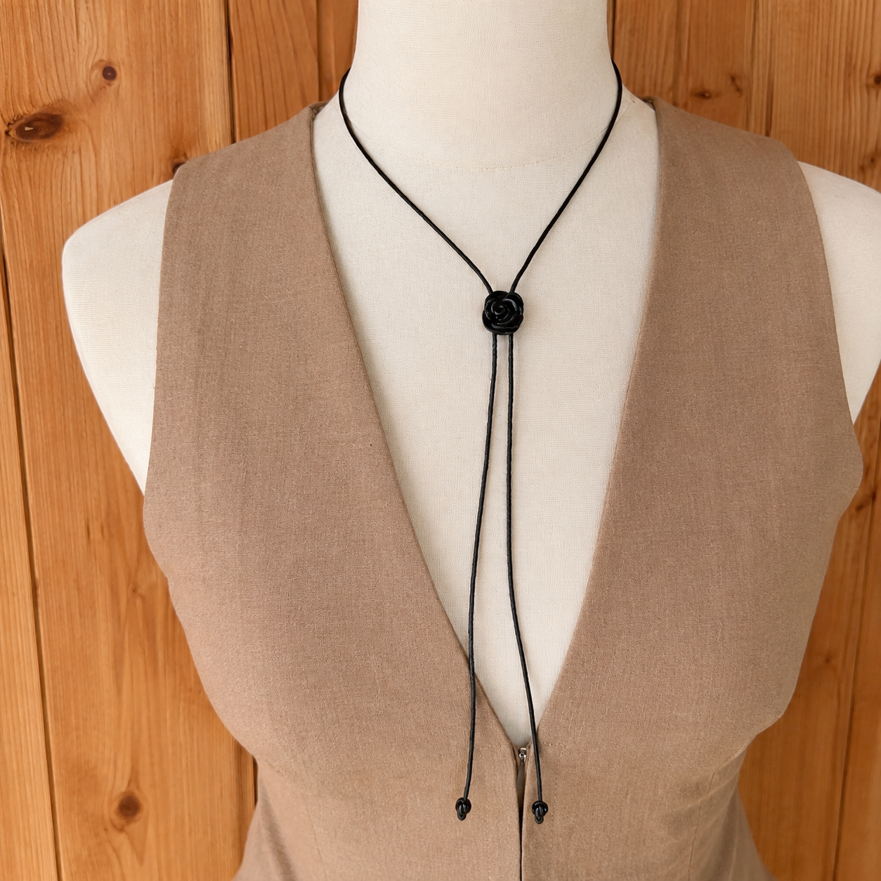 A black cord lariat-style necklace with a round black button closure, displayed on a white mannequin torso wearing a beige sleeveless top, set against a wooden plank background.