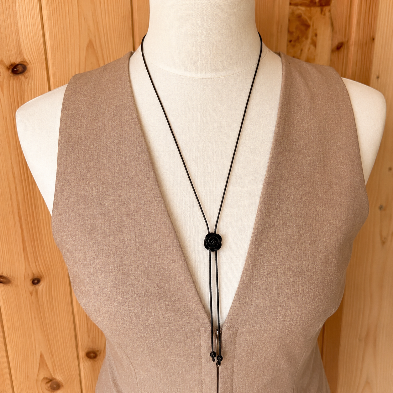 A black cord lariat necklace with a round black bead pendant displayed on a white mannequin torso wearing a beige sleeveless top, set against a light wood panel background.