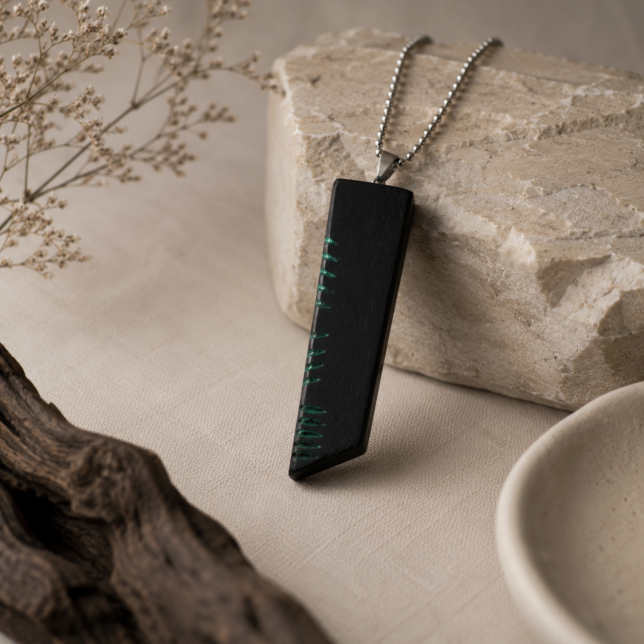 Black pendant with green accents on a chain, placed on a textured surface with natural elements.