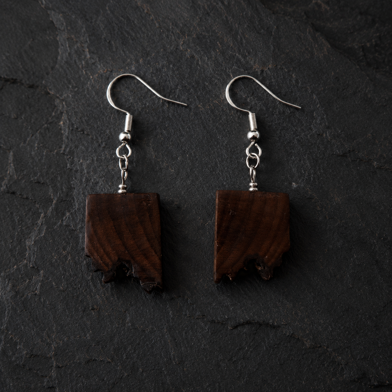 A pair of handmade dangle earrings featuring rectangular dark brown wood pendants with natural rough edges, suspended from silver fishhook ear wires, displayed on a dark textured slate background.