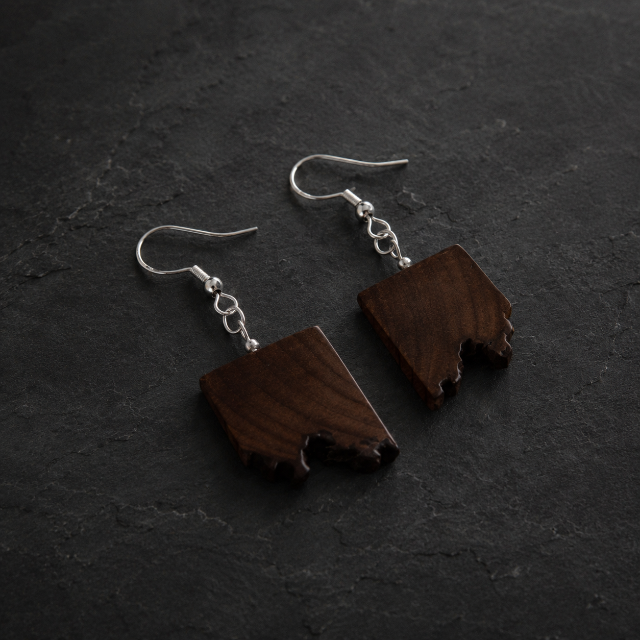 A pair of handmade dangle earrings featuring irregularly shaped dark brown wood pendants with natural rough edges, suspended from silver fishhook wires, displayed on a dark textured slate surface.