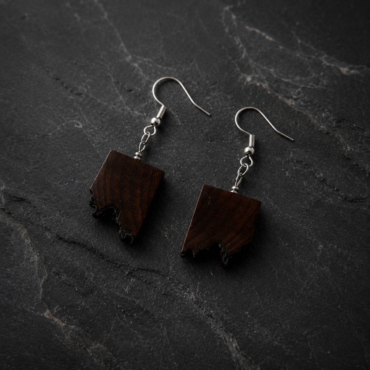 A pair of handmade dangle earrings featuring irregularly shaped dark brown wooden pendants with natural rough edges, suspended from silver fishhook ear wires, displayed on a dark textured slate surface.