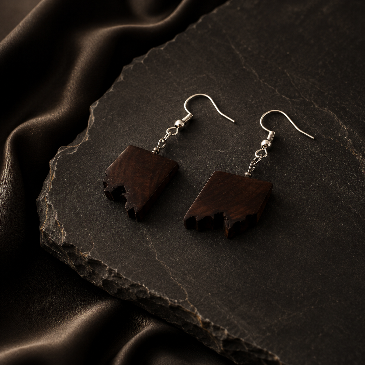 A pair of dark brown wooden dangle earrings with silver fishhook wires, displayed on a dark textured slate slab with draped brown fabric.