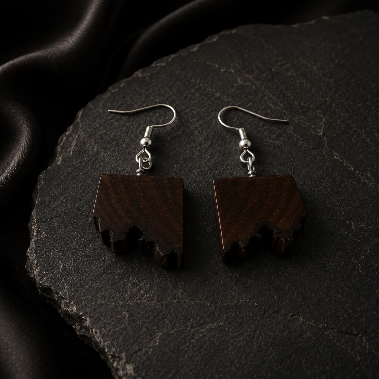 A pair of drop earrings with dark brown wood pendants shaped like stylized leaves, featuring silver-tone fishhook wires, displayed on a dark textured stone surface.