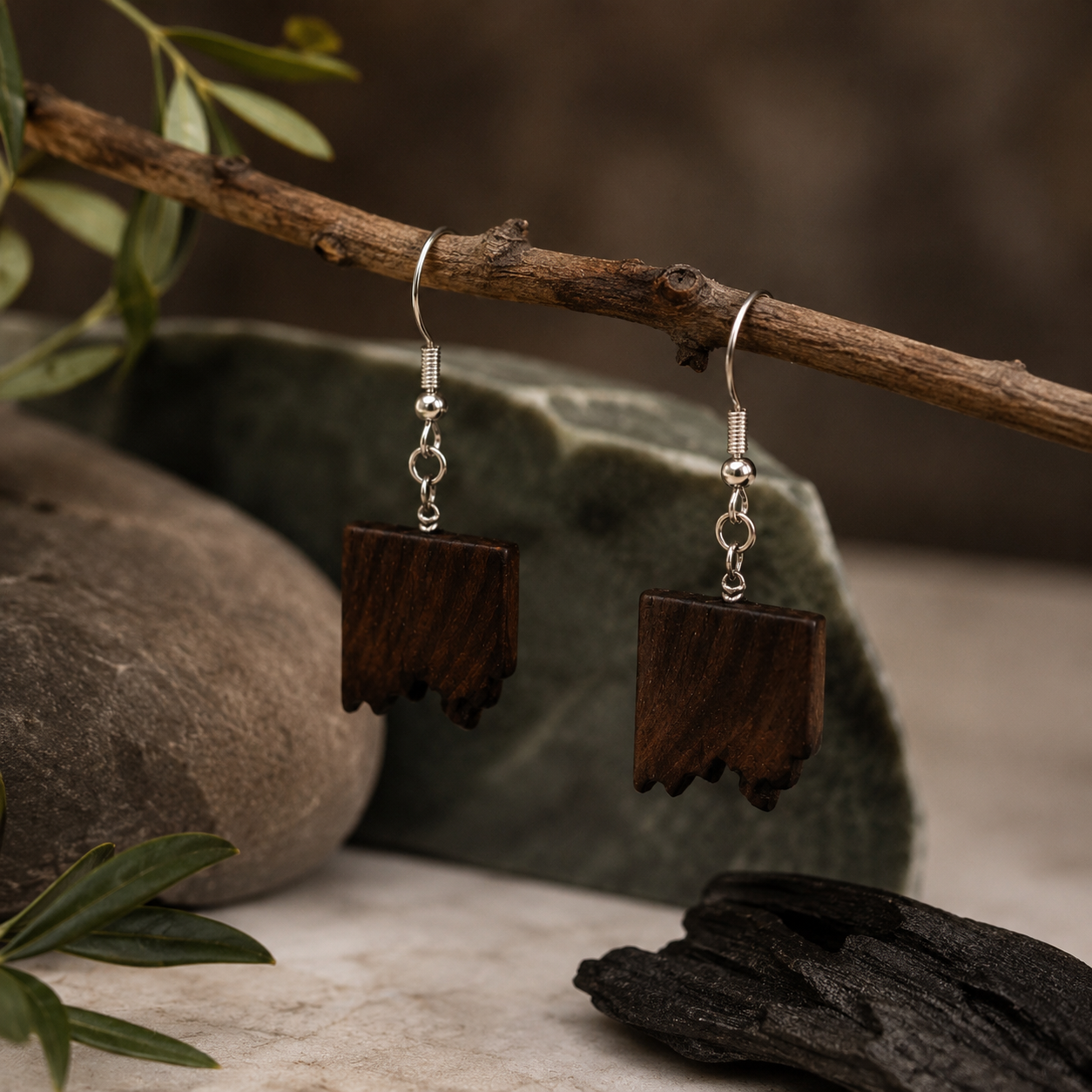 A pair of handmade wooden dangle earrings with silver fishhook wires, hanging from a rustic wooden branch. The earrings feature rectangular dark brown wood pendants with a rustic, melted-edge bottom.