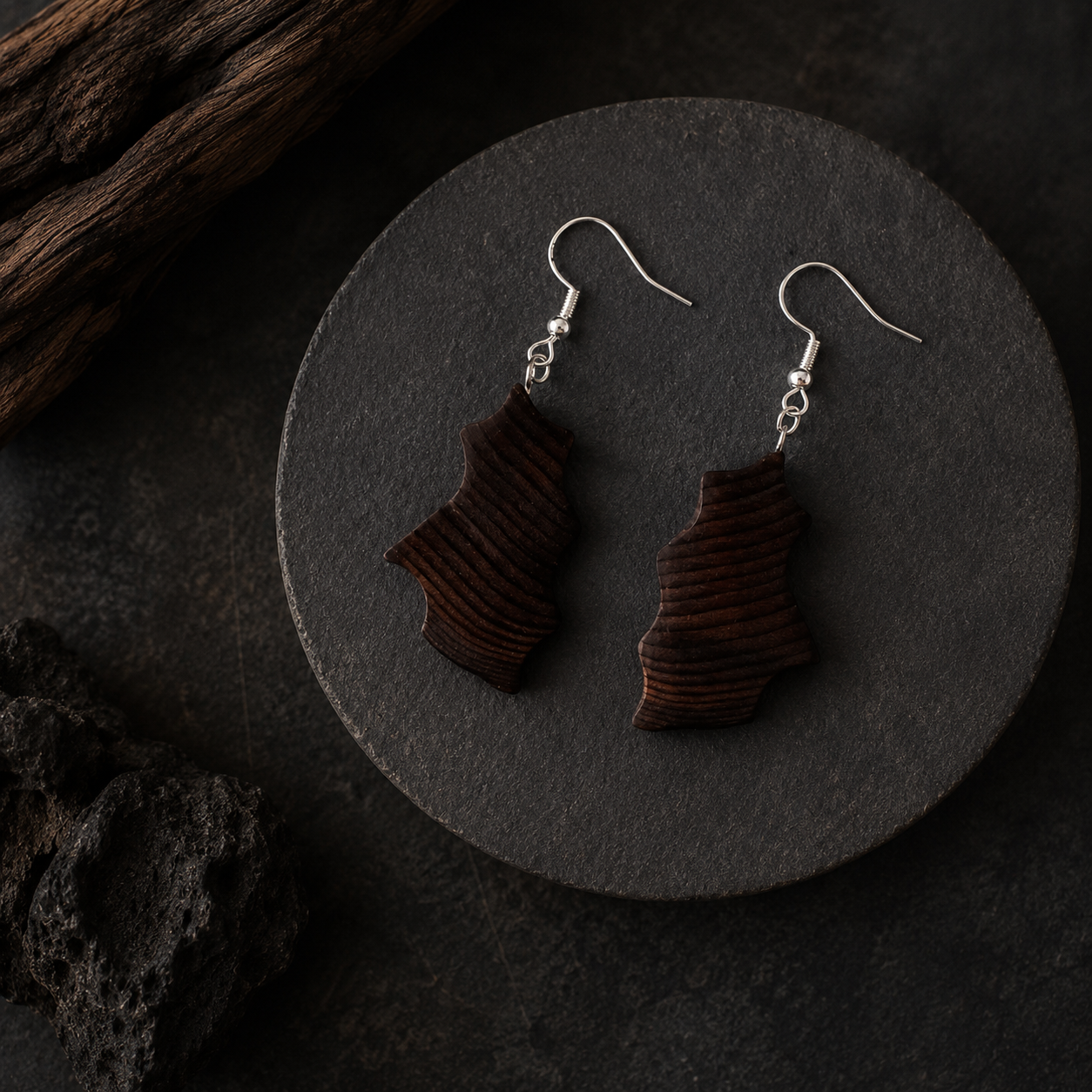 A pair of dark brown wooden drop earrings with a ribbed texture and silver fishhook wires, displayed on a round black stone plate.