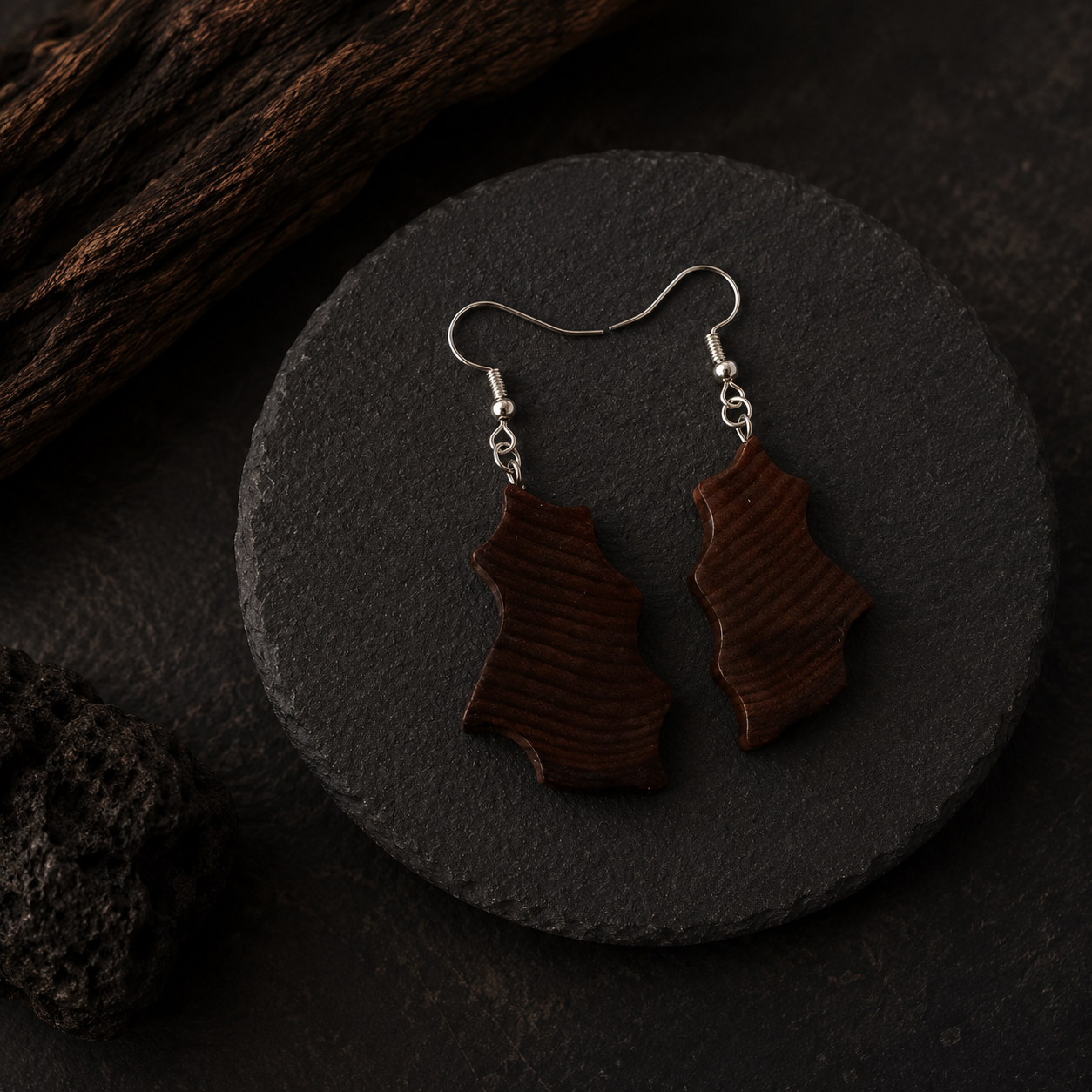 A pair of dark brown wooden drop earrings with silver fishhook wires, displayed on a round black slate coaster.