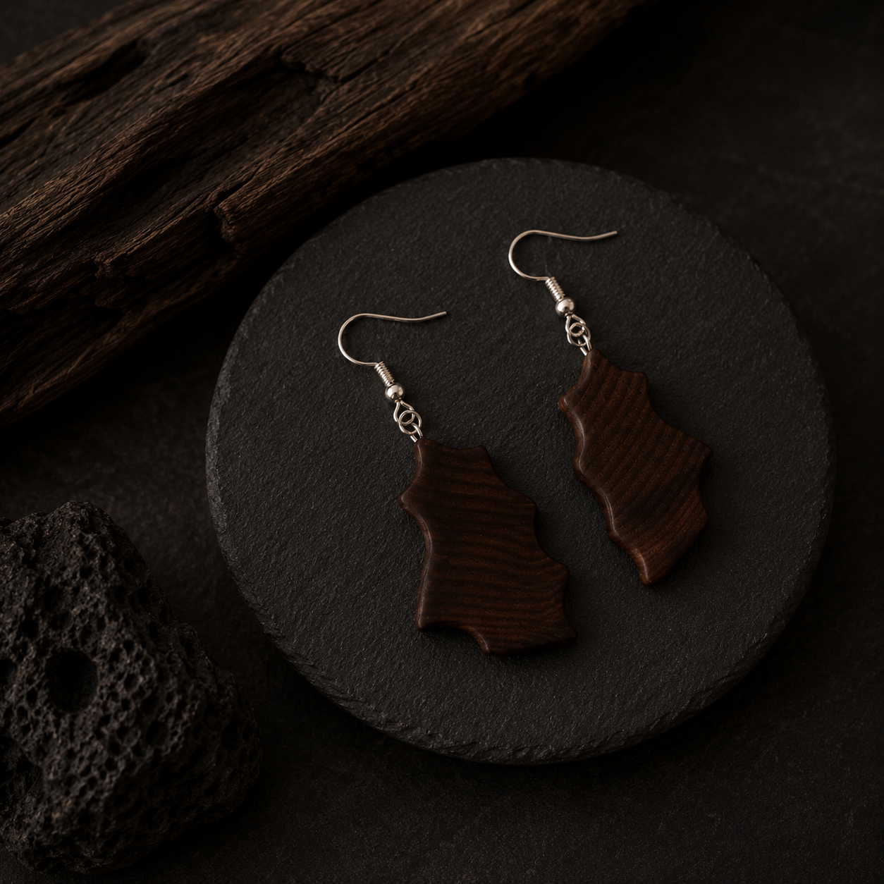 A pair of dark brown wooden drop earrings with silver fishhook wires, displayed on a round black slate coaster with rustic wood and a lava stone in the background.