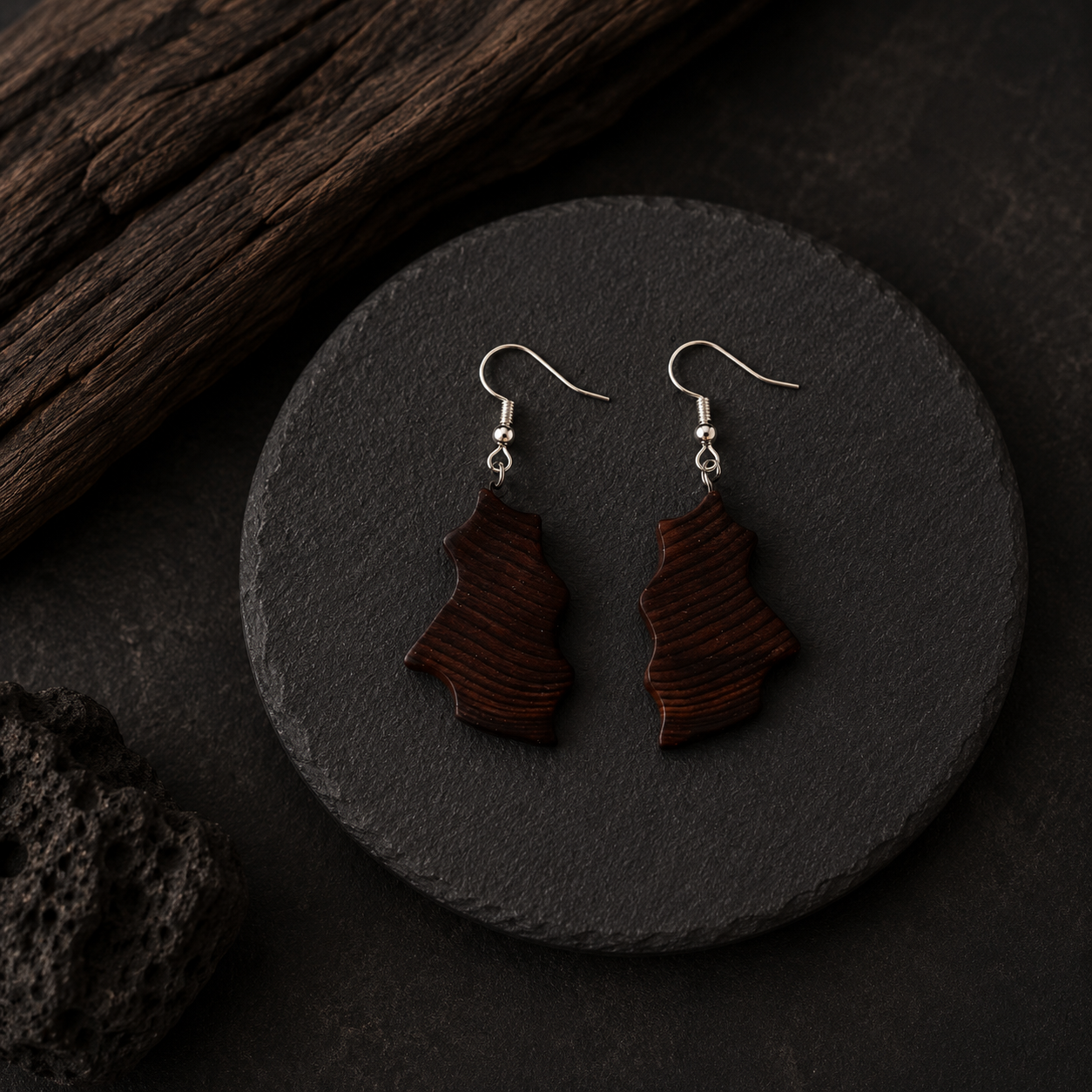 A pair of dark brown wooden dangle earrings with a wavy, layered organic shape and silver fishhook ear wires, displayed on a round black slate coaster.