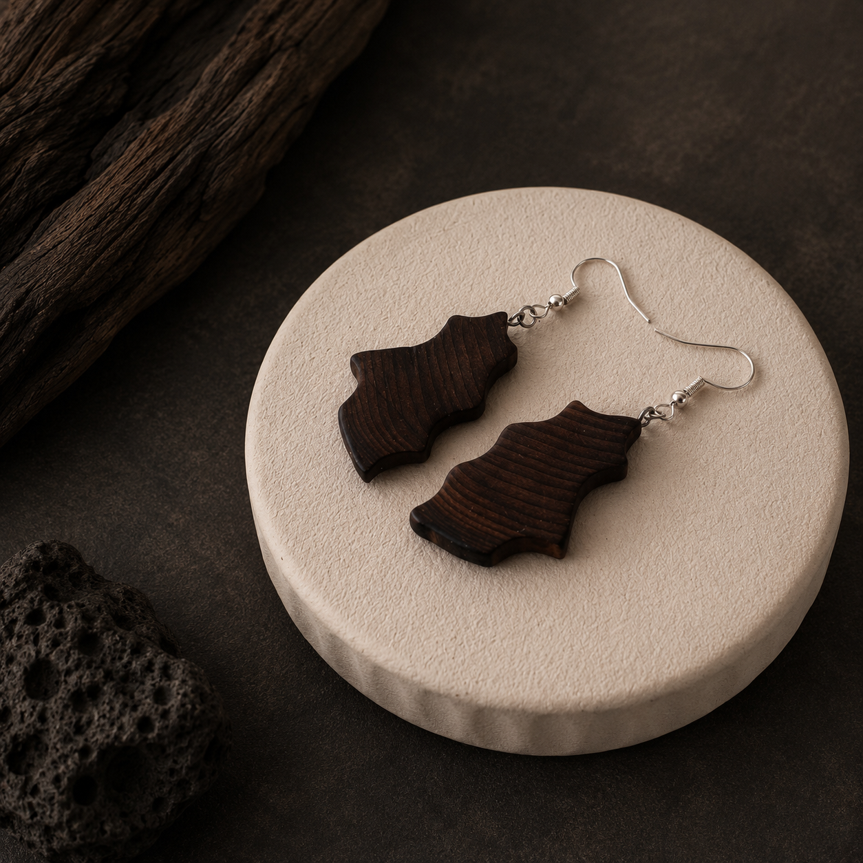 A pair of dark brown wooden dangle earrings with silver fishhook wires, displayed on a round beige ceramic plate against a dark textured background with rustic wood props.