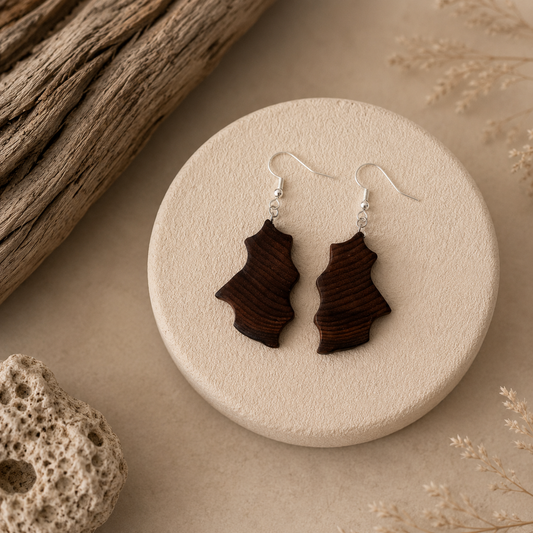 A pair of dark brown wooden dangle earrings with silver fishhook wires, displayed on a round beige textured stand with rustic wood and dried flowers in the background.