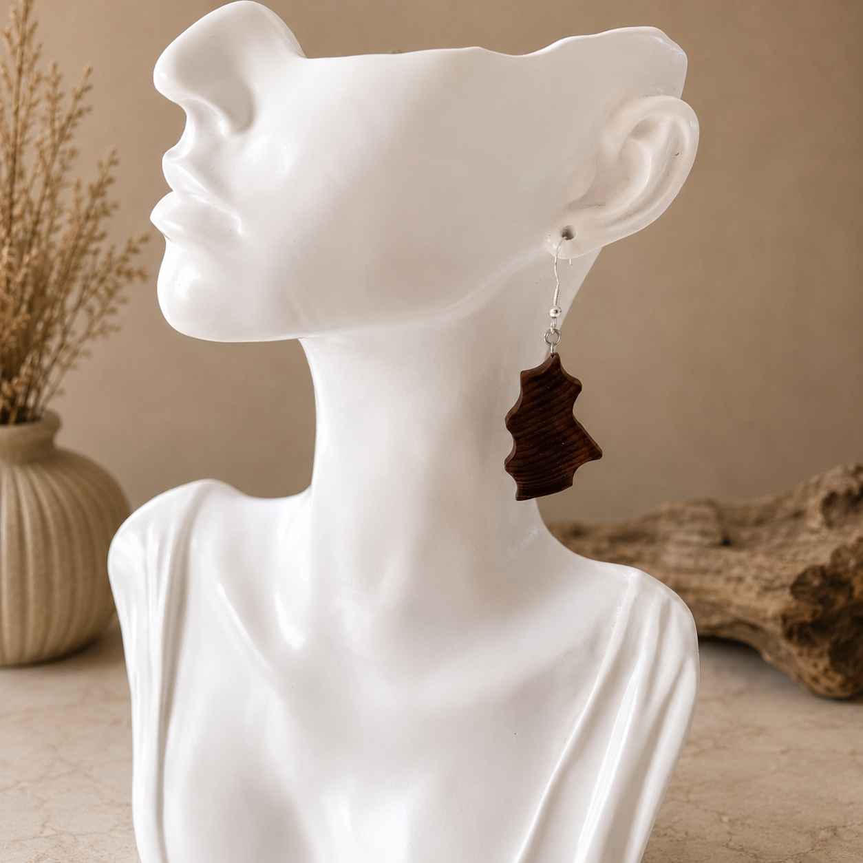 A dark brown wooden dangle earring with a silver-toned post, shown hanging from the ear of a white ceramic female bust. The background features a warm beige wall, a dried plant in a vase, and a piece of driftwood.