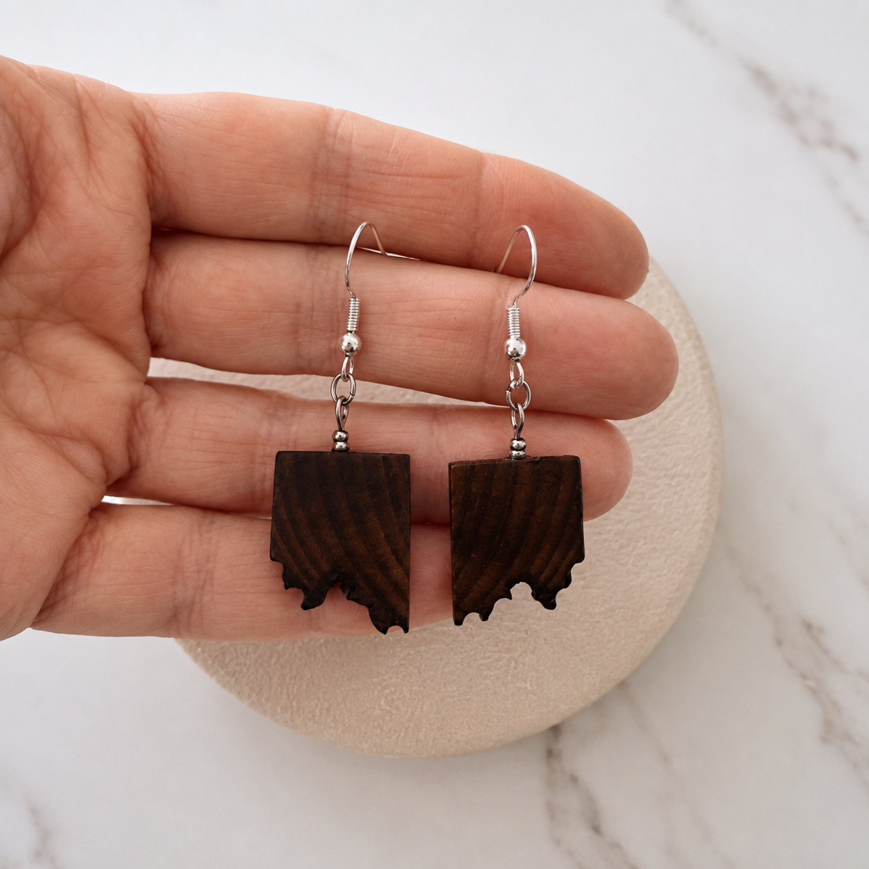 A pair of dark brown wood drop earrings shaped like the state of Idaho, held in a person's hand against a light marble background.