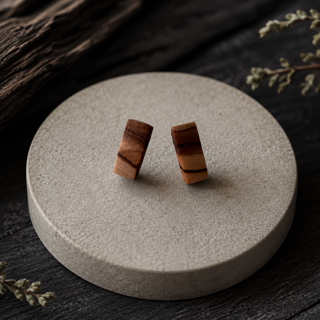 A pair of small wooden post earrings with natural brown and tan grain, displayed on a round textured beige stone coaster over a dark wood surface.