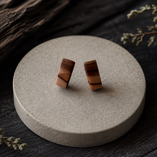 A pair of small wooden post earrings with natural brown and tan grain, displayed on a round textured beige stone coaster over a dark wood surface.