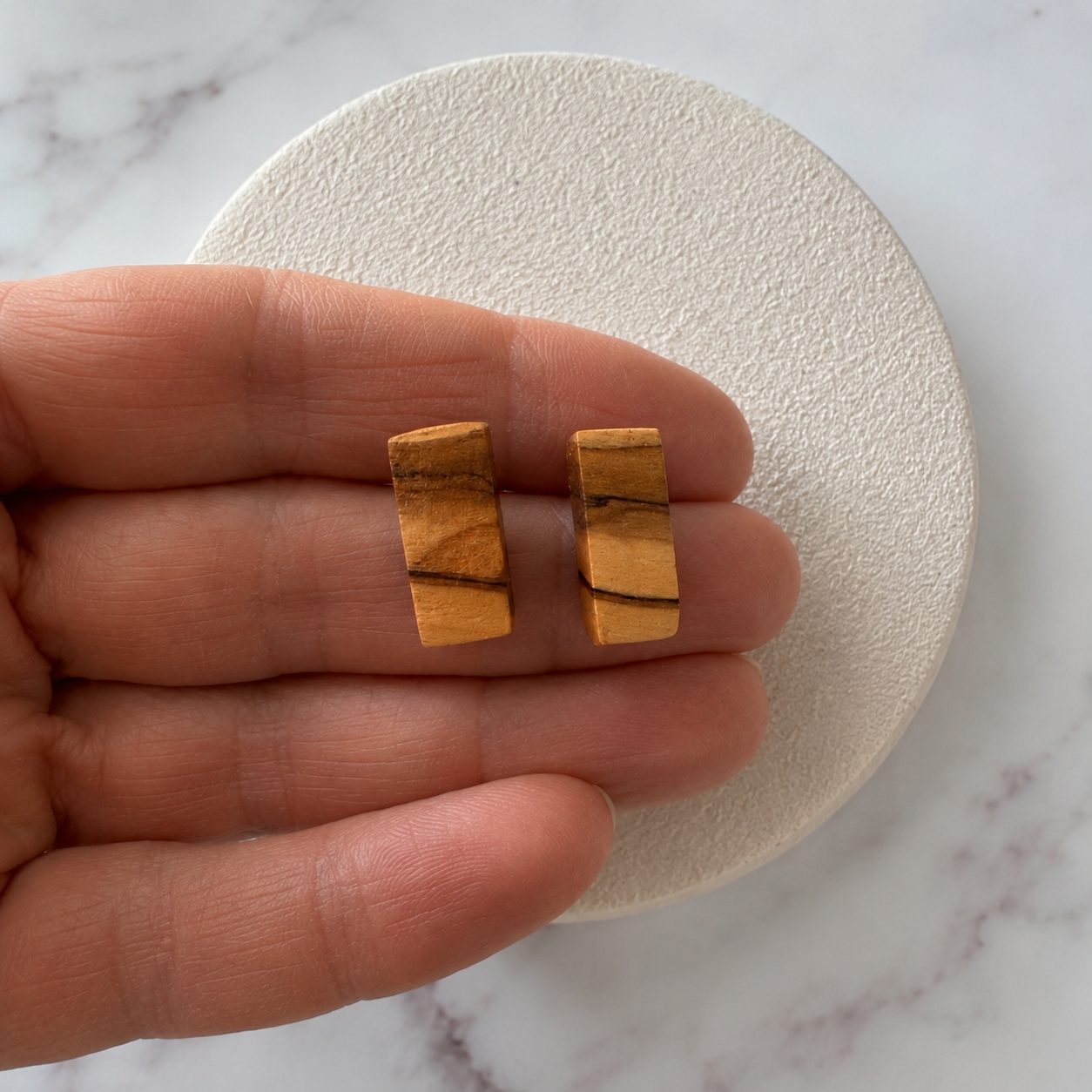 A pair of small wooden rectangular stud earrings with natural wood grain, held between a person's fingers over a textured white circular display on a marble surface.