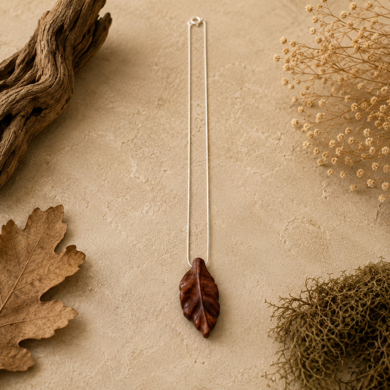 Silver necklace with a leaf pendant on a beige surface with natural elements.