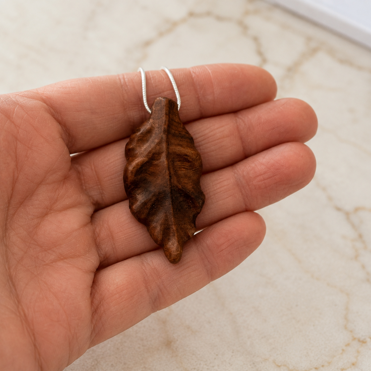 Hand holding a brown leaf-shaped pendant against a marble background